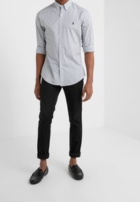 Mężczyzna w jasnej, w paseczki koszuli z podwiniętymi rękawami, czarnych, wywiniętych spodniach oraz czarnych loafersach stoi na białym tle.