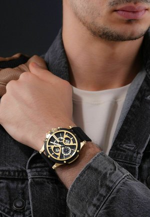 Homme portant une montre-bracelet noire et dorée à grand cadran avec plusieurs cadrans, habillé d'une chemise blanche et d'une veste en denim foncé, tenant un sac marron.
