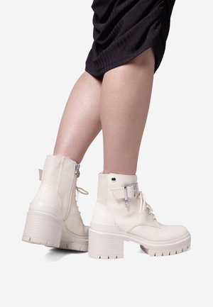 Stivali alla caviglia chunky in pelle bianca, con zip laterale, apertura con lacci e un accentuato cinturino. Tacco spesso e testurizzato con suola scolpita.