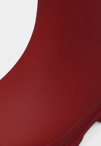 Κοντινό πλάνο λείας, γυαλιστερής κόκκινης λαστιχένιας μπότες βροχής που δείχνει τον αστράγαλο και μέρος της σόλας σε απλό λευκό φόντο.