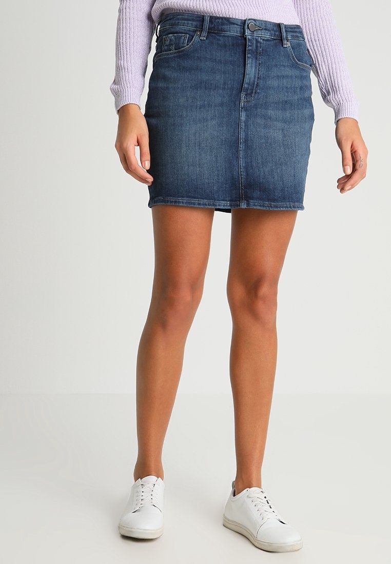 Jeansowa mini spódnica w ciemnoniebieskim kolorze o dopasowanym kroju, z zapięciem na guziki z przodu i pięcioma kieszeniami. Nosi się z białymi trampkami.