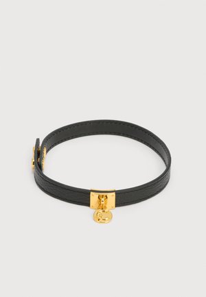 Collana choker in pelle nera con chiusura quadrata color oro e piccolo ciondolo rotondo inciso con la lettera "C" su uno sfondo bianco.