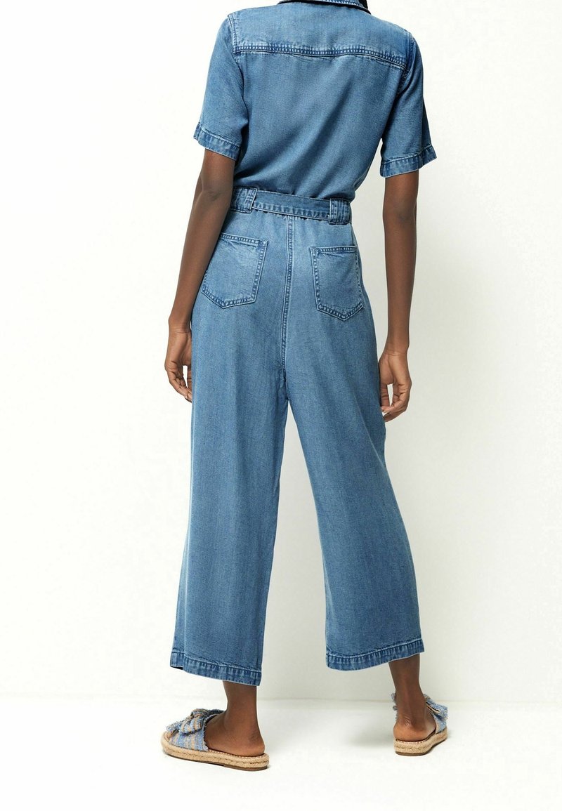 Personne portant une combinaison en denim bleu clair à manches courtes et jambes larges, debout pieds nus sur un fond blanc.