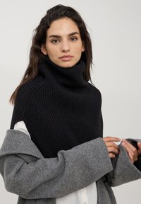 Maglione nero a coste con colletto alto e maniche corte, abbinato a un cappotto di lana grigio con maniche ampie e dettaglio dei bottoni.
