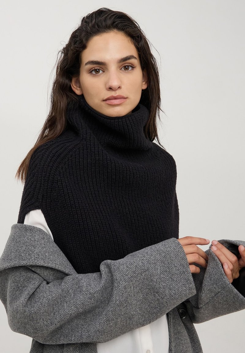 Maglione nero a coste con colletto alto e maniche corte, abbinato a un cappotto di lana grigio con maniche ampie e dettaglio dei bottoni.