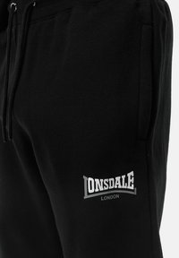 Zwarte katoenen joggers met een elastische tailleband, voorzien van een witte "Lonsdale London" logo op de linker dij. Zachte textuur, casual ontwerp.