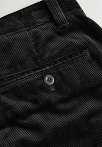 Black corduroy fabric with vertical ribs, featuring a single round button near the waistband and a back pocket detail.