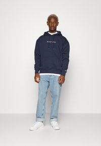 Sudadera azul marino con logo blanco, bolsillo frontal y cordón; combinada con jeans azul claro y zapatillas blancas. Diseño simple y casual.