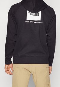Persona que lleva una sudadera negra con capucha del logo de The North Face y el texto "Never Stop Exploring" en la parte de atrás, combinada con pantalones beige.