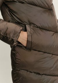 Smeđi puffer kaput s sjajnom završnom obradom, s širokim kvadratnim dijelovima, bočnom džepom i kopčama na rukavima.