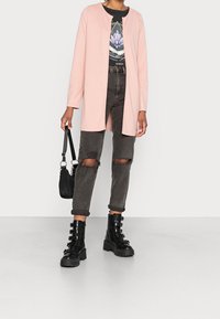 Cardigan lungo rosa chiaro sopra una t-shirt grafica nera, abbinato a jeans scuri strappati e stivaletti neri chunk. Borsa nera a tracolla.