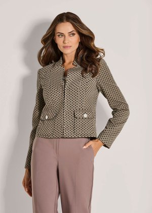 Frau mit langen welligen Haaren, die eine strukturierte braune Jacke mit Muster und Fronttaschen sowie mauve hoch taillierte Hose trägt, Hand in der Tasche.