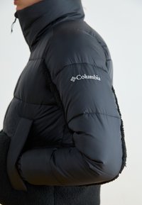 Svart pufferjakke med høy krage, quiltet tekstur og fleece-detaljer. Columbia-logo brodert på høyre arm.