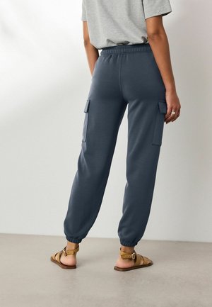 Persoană purtând pantaloni cargo jogger bleumarin și sandale maro cu bretea la gleznă, stând cu spatele la cameră pe un fundal simplu.