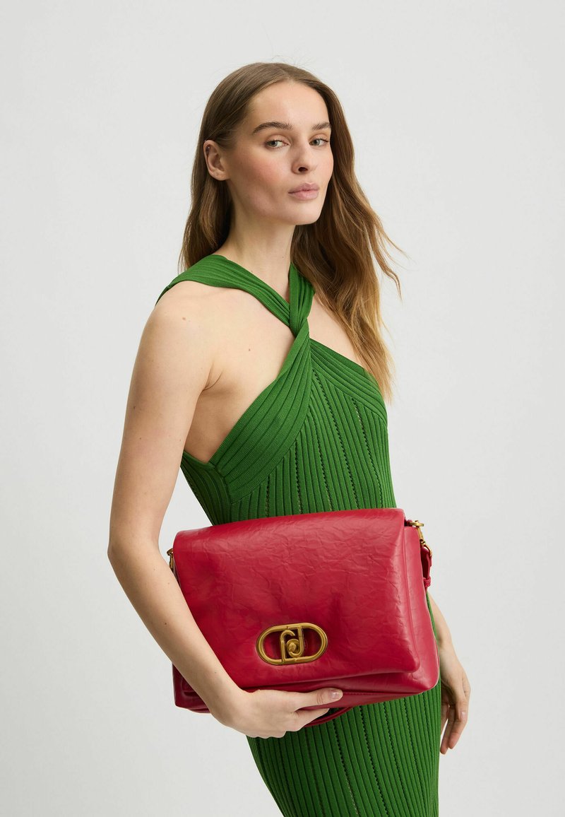 Pochette en cuir rouge avec une texture froissée, des accessoires en métal doré, et une forme rectangulaire, tenue contre une robe côtelée verte.