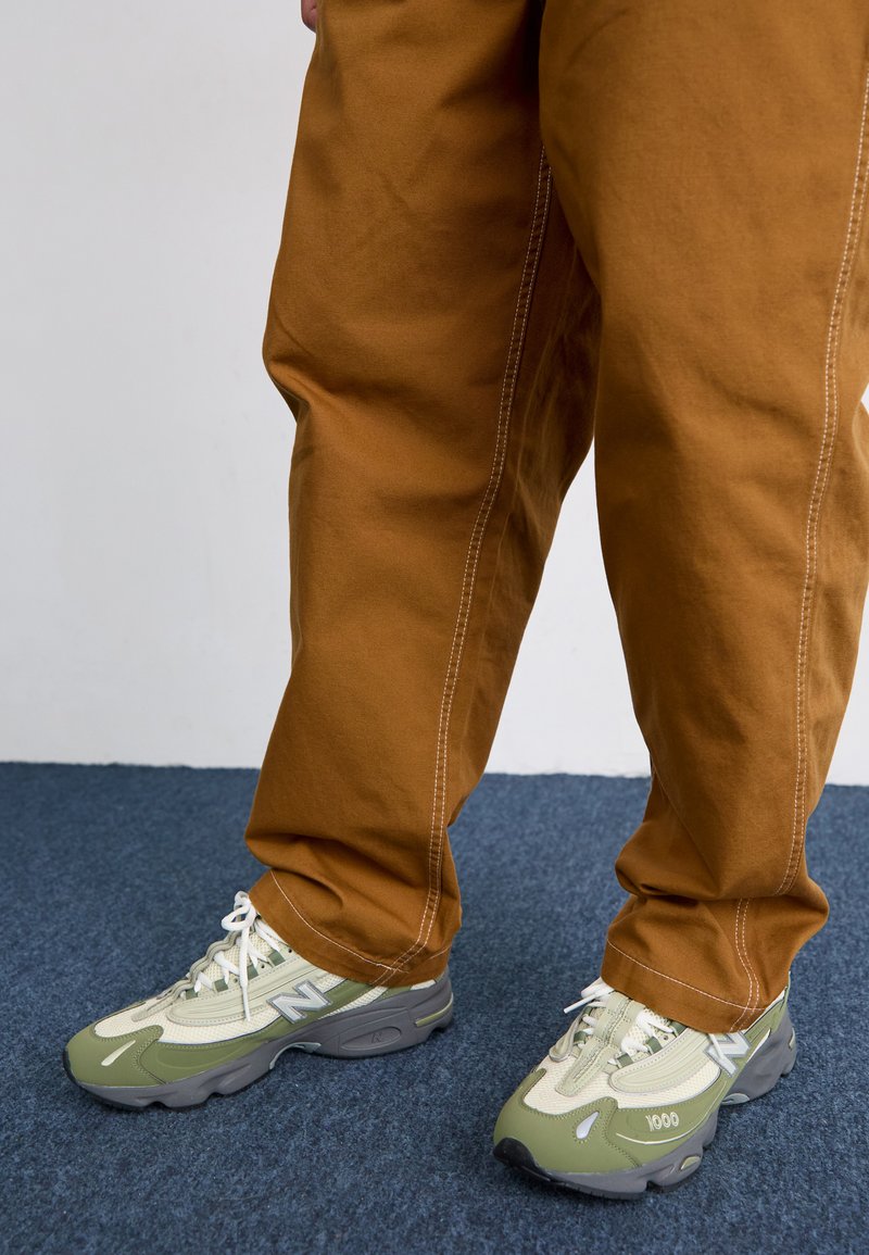 Zapatillas deportivas de color verde claro y gris, con materiales de malla y sintéticos, presentan cordones blancos y el texto "1000" en el lateral. Pantalones marrones con costuras blancas.