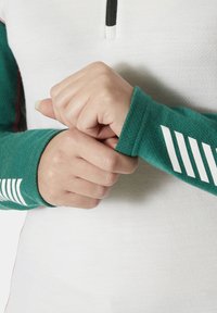 Zielona bluzka z długim rękawem, z zamkiem błyskawicznym i teksturowanym białym przodem. Lewy rękaw ozdobiony jest białymi ukośnymi paskami. Materiał wydaje się miękki i elastyczny.