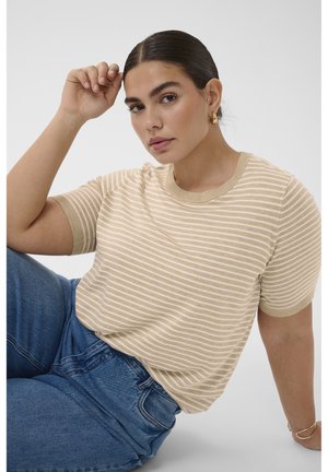 Femme aux cheveux foncés portant un haut beige et blanc à manches courtes rayé et un jean bleu, assise avec un bras levé près de la tête.