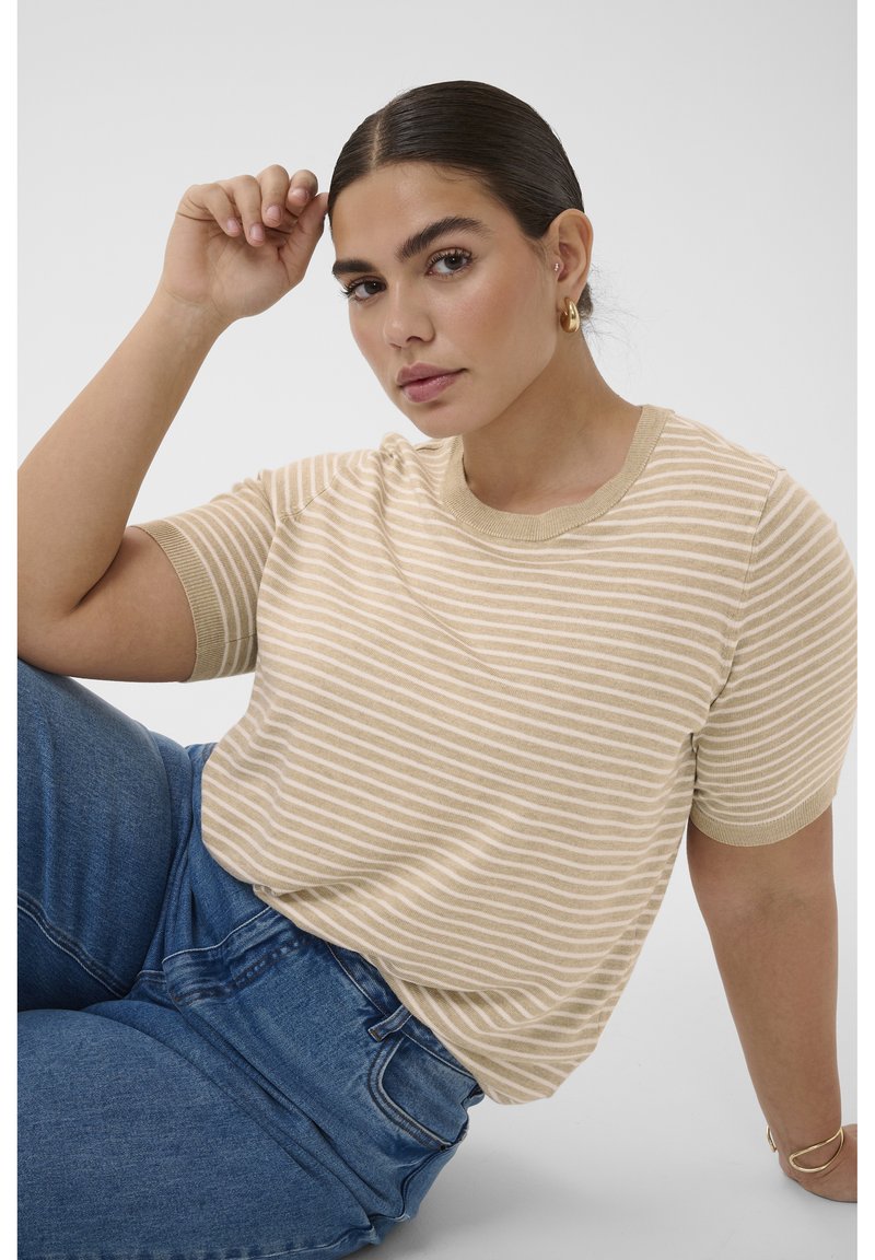 Frau mit dunklen Haaren, die ein beige-weiß gestreiftes kurzärmliges Oberteil und blaue Jeans trägt, sitzt mit einem Arm erhoben nahe ihrem Kopf.