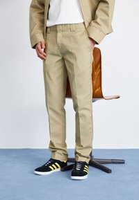 Beige skreddersydde bukser med rett snitt, laget av bomullsstoff. Kombinert med svarte joggesko med gule striper.