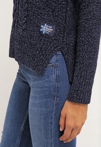 Stickad tröja i mörkblått, med ett strukturerat mönster, sidospår och en märkespatch med "Knit Super Dry Co." nederst till vänster.