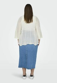 Weißer Strickcardigan mit Durchbrüchen, drei Viertel Glockenärmeln, kombiniert mit einem blauen Jeans-Midi-Rock. Weiße Slipper mit schwarzen Akzenten.