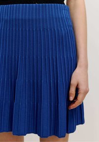 Jupe plissée bleu en tissu tricot à côtes, avec une taille mi-haute et un ourlet évasé. Une main tient le tissu sur le côté.