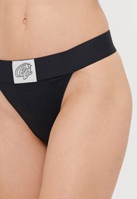 Black thong underwear featuring a wide elastic waistband with a white logo tag. Smooth texture and minimal coverage design.