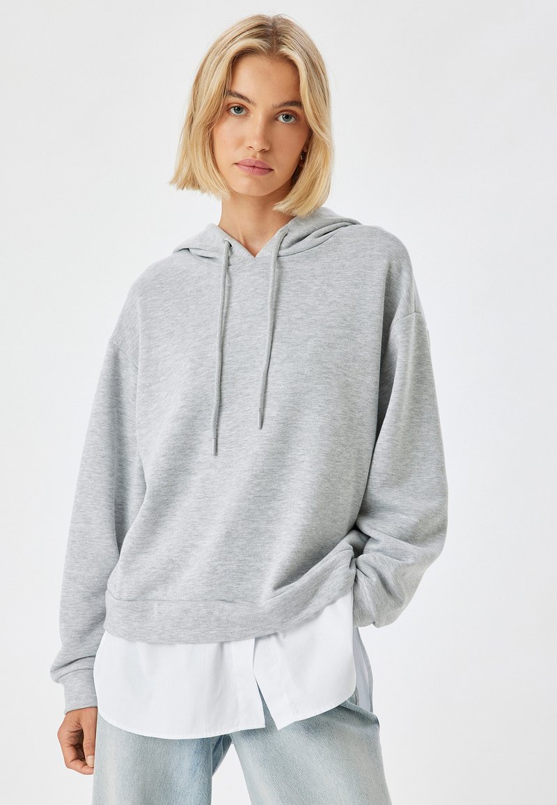 Sudadera gris con manga larga, capucha con cordón y bolsillo frontal tipo canguro, sobre una camiseta blanca. Textura suave, ajuste relajado.