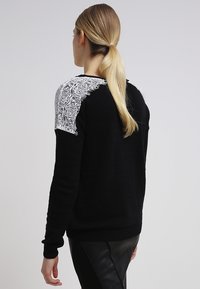 Femme avec queue de cheval blonde portant un pull noir avec un détail en dentelle blanche sur l'épaule et une jupe noire, vue de dos sur un fond uni.