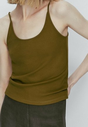 Vrouw die een mouwloos geribbeld olijfgroen tanktopje en donkere broek draagt, getoond van nek tot heup met hand rustend op de heup.