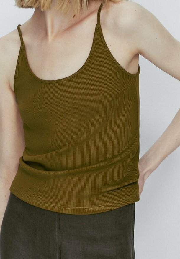Femme portant un débardeur côtelé vert olive sans manches et un pantalon foncé, montrée du cou aux hanches avec la main posée sur la hanche.