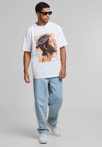 Grafisk hvid t-shirt med et trykt portræt med krone og tekst, parret med lyseblå løstsiddende jeans og hvide sneakers. Sorte solbriller bæres.