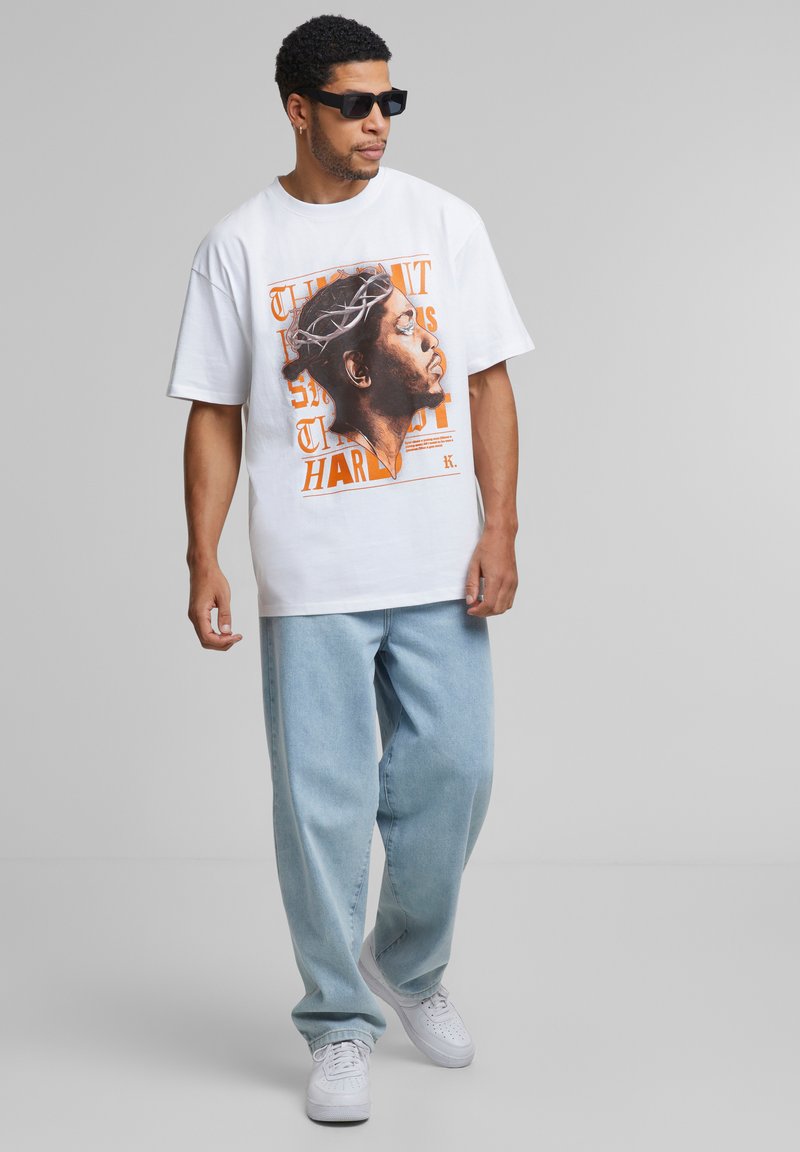 Grafisk hvid t-shirt med et trykt portræt med krone og tekst, parret med lyseblå løstsiddende jeans og hvide sneakers. Sorte solbriller bæres.