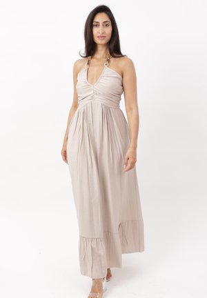 Kvinde iført en ærmeløs beige maxi-kjole med sammensyet buste og halter-stropper på en ensfarvet hvid baggrund.