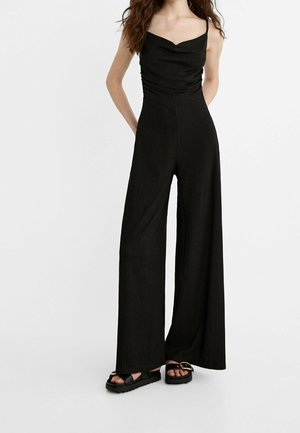 Femme portant une combinaison noire sans manches avec un pantalon à jambes larges et des sandales plateformes noires à boucle dorée sur fond blanc uni.