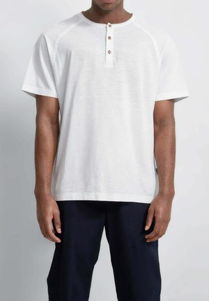 Uomo che indossa una maglietta Henley bianca a maniche corte con tre bottoni e pantaloni scuri, in piedi davanti a uno sfondo chiaro e uniforme.