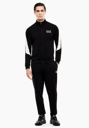 Mand stående iført sort EA7 træningsdragt med hvide striber på ærmerne og sorte EA7 sneakers på hvid baggrund.