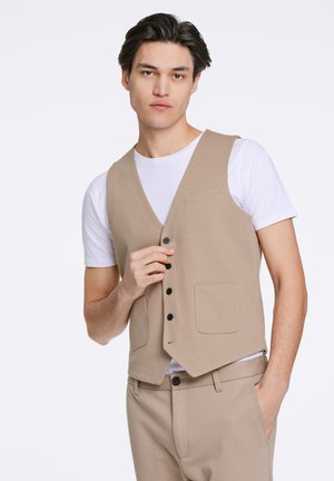 Gilet beige realizzato in tessuto testurizzato, con scollatura a V profonda, cinque bottoni neri e due tasche frontali, indossato sopra una maglietta bianca.