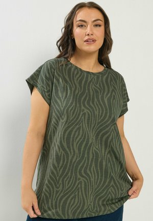 Kvinde iført en grøn kortærmet top med et bølget, zebra-lignende mønster, stående mod en ensfarvet lys væg.