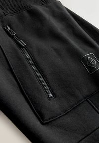 Schwarze Jogginghose mit einem strukturierten Bund, ausgestattet mit einer seitlichen Reißverschlusstasche, flachen Nähten und einem kleinen Logopatch. Glattes Material mit minimalen Details.