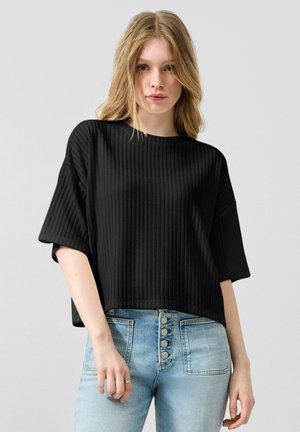 Jeune femme aux cheveux blonds portant un haut noir côtelé à manches courtes et un jean taille haute bleu clair avec des boutons.