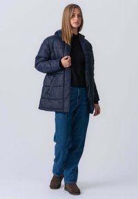 Marineblå pufferjakke med hætte, der har et quiltemønster, frontlynlås og sidelommer, parret med mørkeblå jeans og brune støvler.