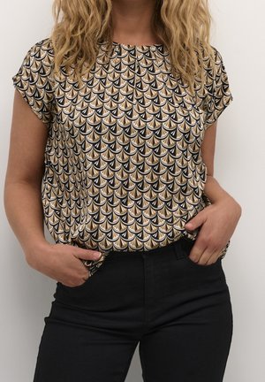 Femme portant un haut à manches courtes avec un motif éventail noir, beige et blanc, rentré dans un pantalon noir taille haute, mains dans les poches.