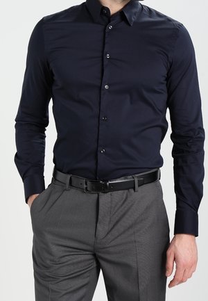 Man draagt een nauwsluitend donkerblauw overhemd met knopen, ingestopt in grijze nette broek met een zwarte leren riem, handen gedeeltelijk in de zakken.