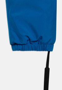 Blue elastic cuff of a garment featuring a gathered texture, with a black fabric tag protruding from the side for brand identification.