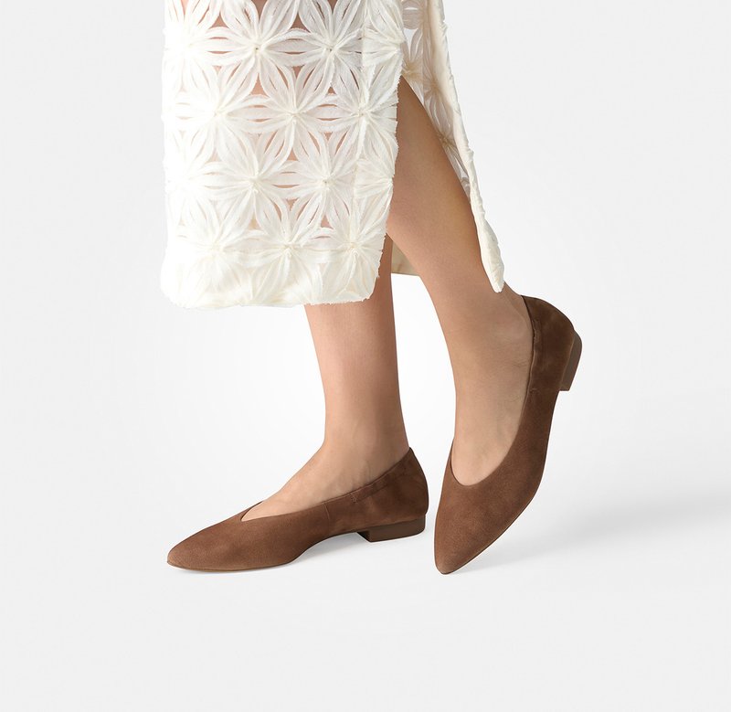 Ben i brune spidse flade sko kombineret med en hvid, blomstret tekstureret midi-nederdel med sideslids på hvid baggrund.