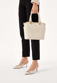 Biała skórzana torebka ze złotymi okuciami, o usztywnionym kształcie, z małymi bocznymi kieszeniami i górnym uchwytem. Do tego beżowe sandały na obcasie typu slingback z ozdobnymi detalami.