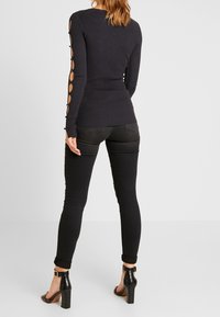 Femme portant un pull noir ajusté avec des manches découpées à boutons, un jean skinny noir retroussé et des sandales à talons carrés noires, debout.