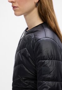 Profil d'une femme aux longs cheveux portant des boucles d'oreilles anneaux dorées et une veste matelassée noire avec un petit patch logo sur la manche.